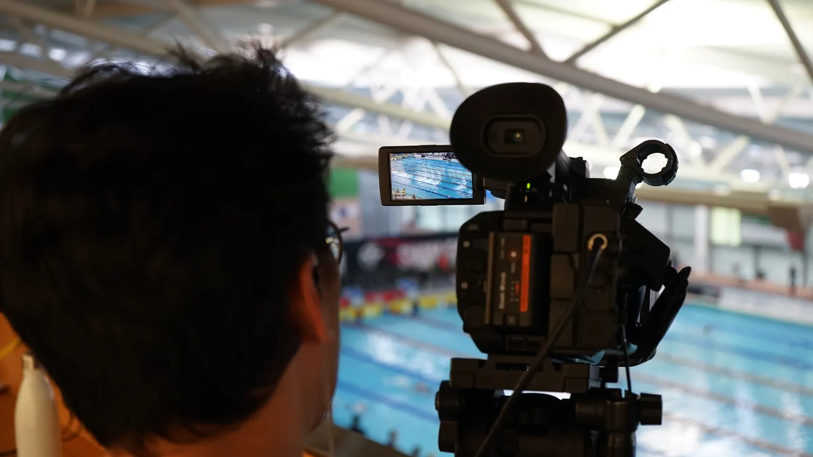 An ARCAE SPORTS camera operator covering a race, the swimmers visible on the camera monitor with the competition pool behind.
