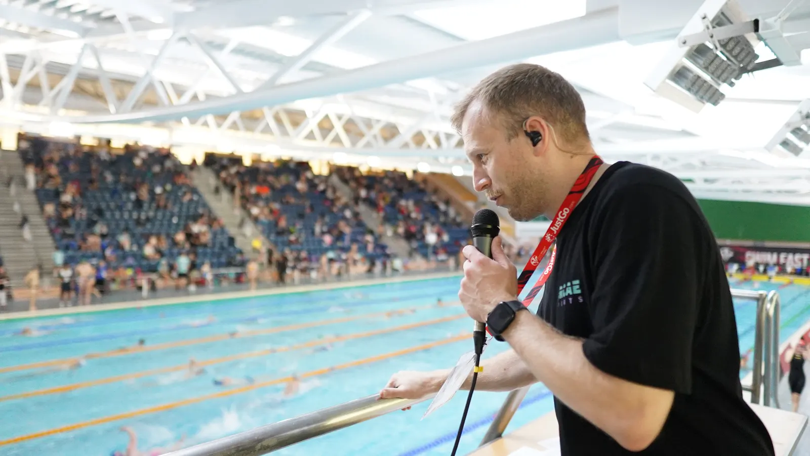 An ARCAE SPORTS commentator on microphone at poolside, in front of a full spectator stand, as a race runs in the pool below.